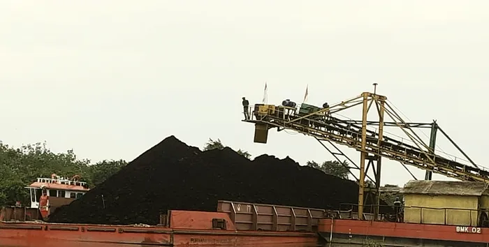 Port stockpile of coal beside moored barges with a loading conveyor overhead.