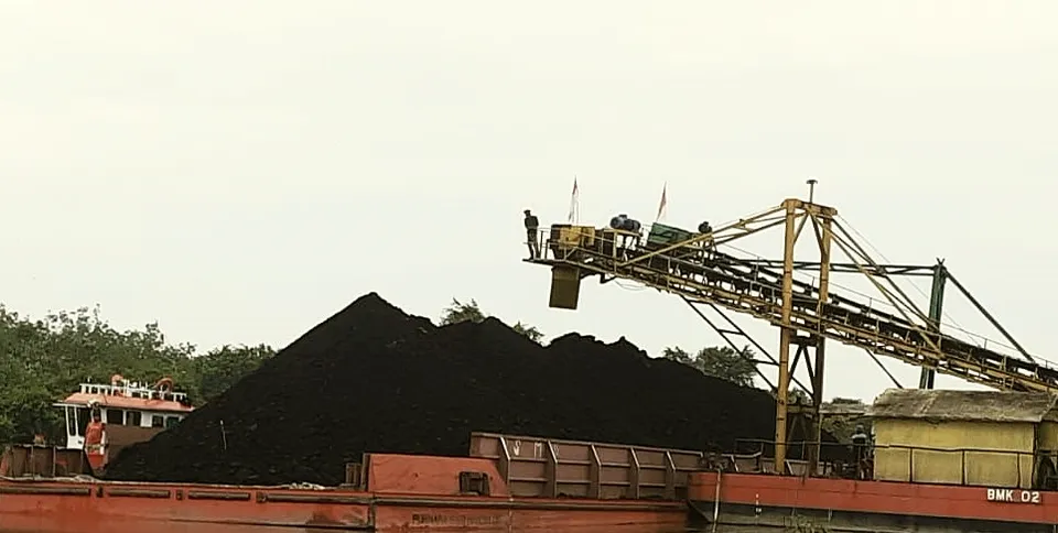 Coal stockpile beside moored barges at a river port, loading gantry overhead.