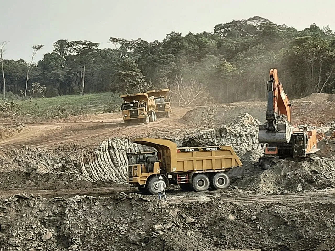 Excavator and haul trucks working an overburden face with forest on the ridge above.
